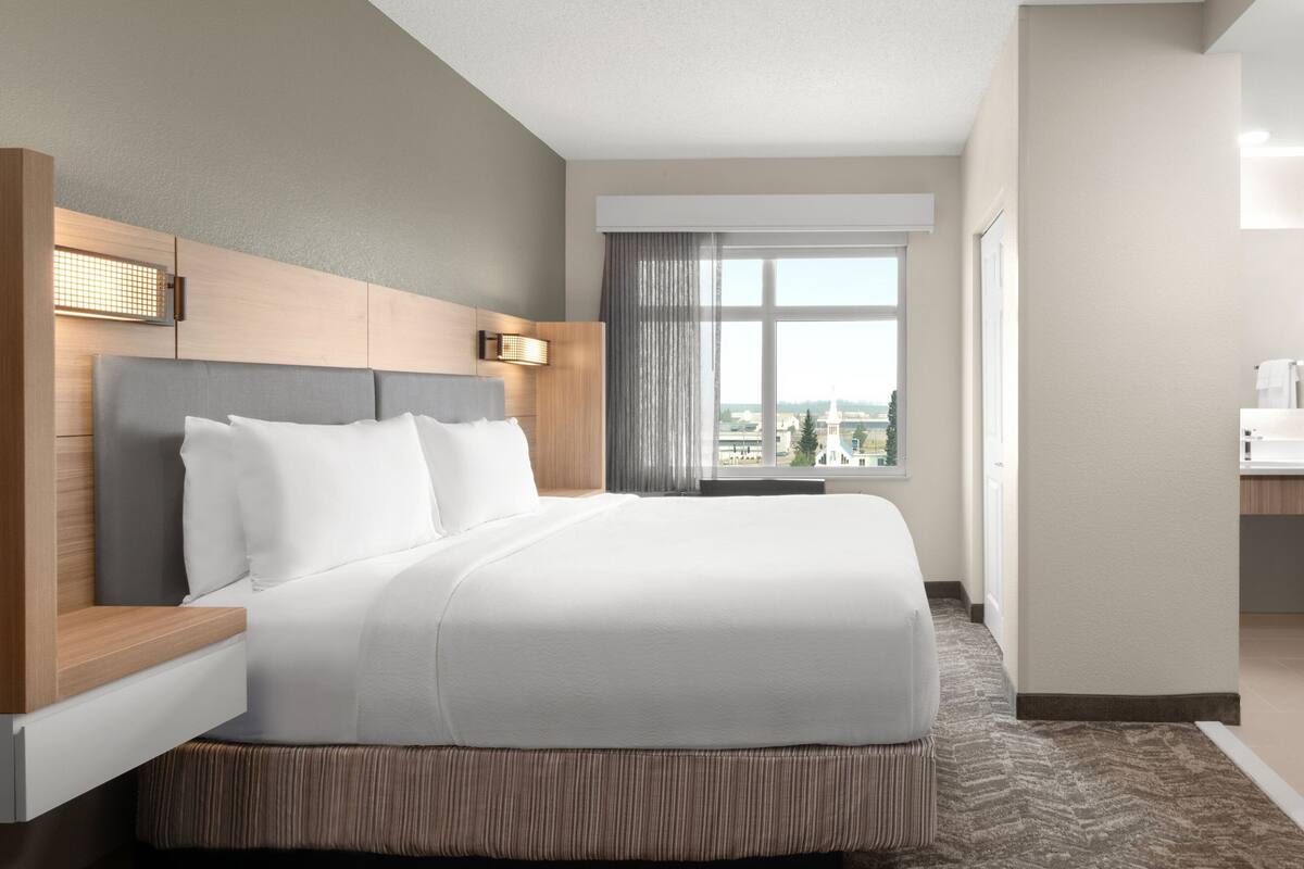 A hotel room with a large bed, a wooden headboard, a nightstand, a window with curtains, and a view of buildings outside.