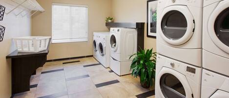 A laundry room with multiple washing machines, a drying rack, a potted plant, and a window with blinds.