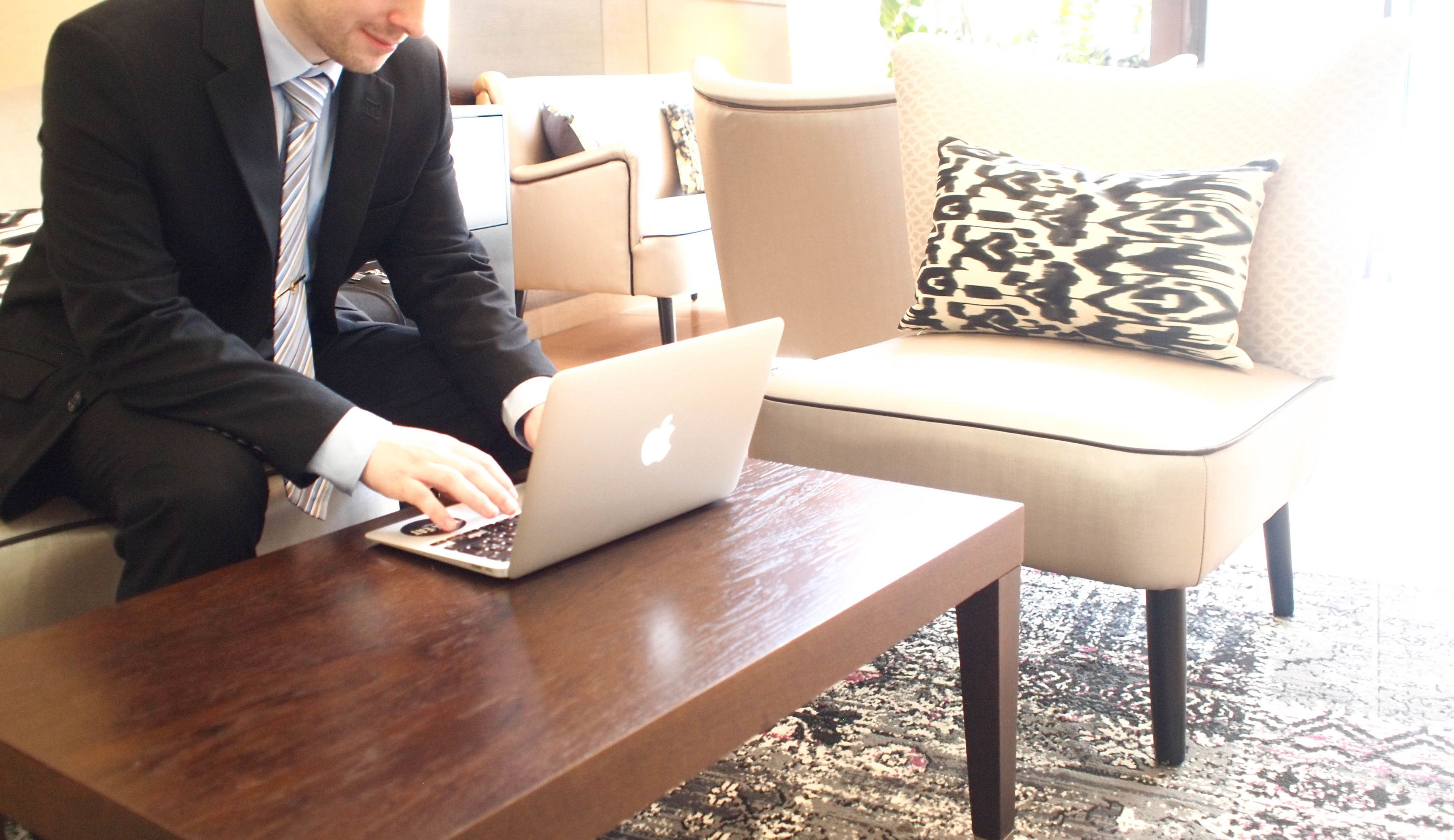 A man in a suit using a laptop on a wooden table in a well-lit room with a sofa and patterned pillows.