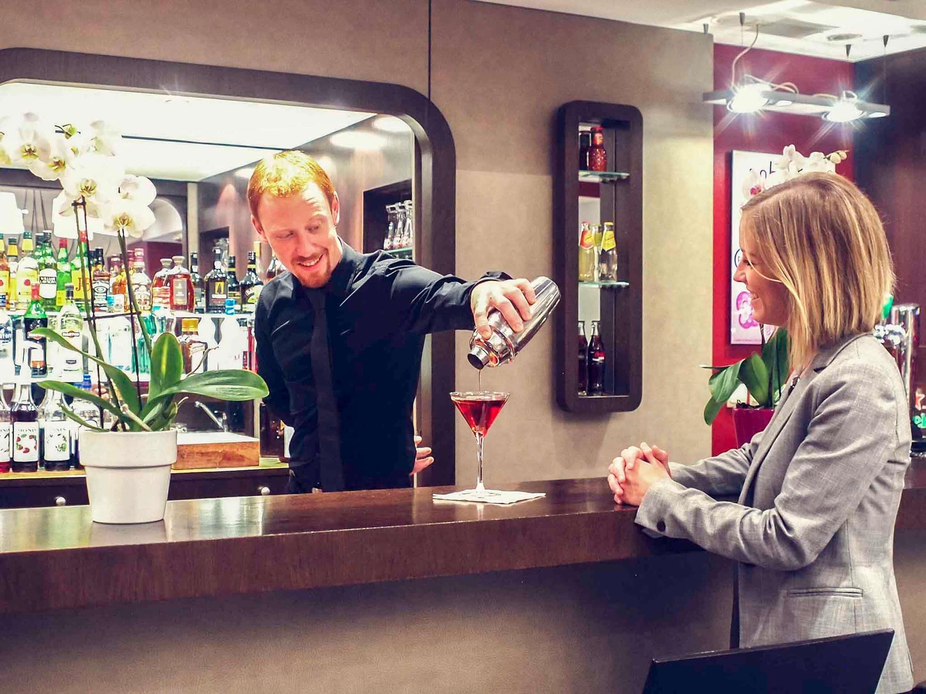Zona de bar con un bartender sirviendo una bebida en una copa de martini, una mujer sentada en la barra, y varias botellas y plantas al fondo.