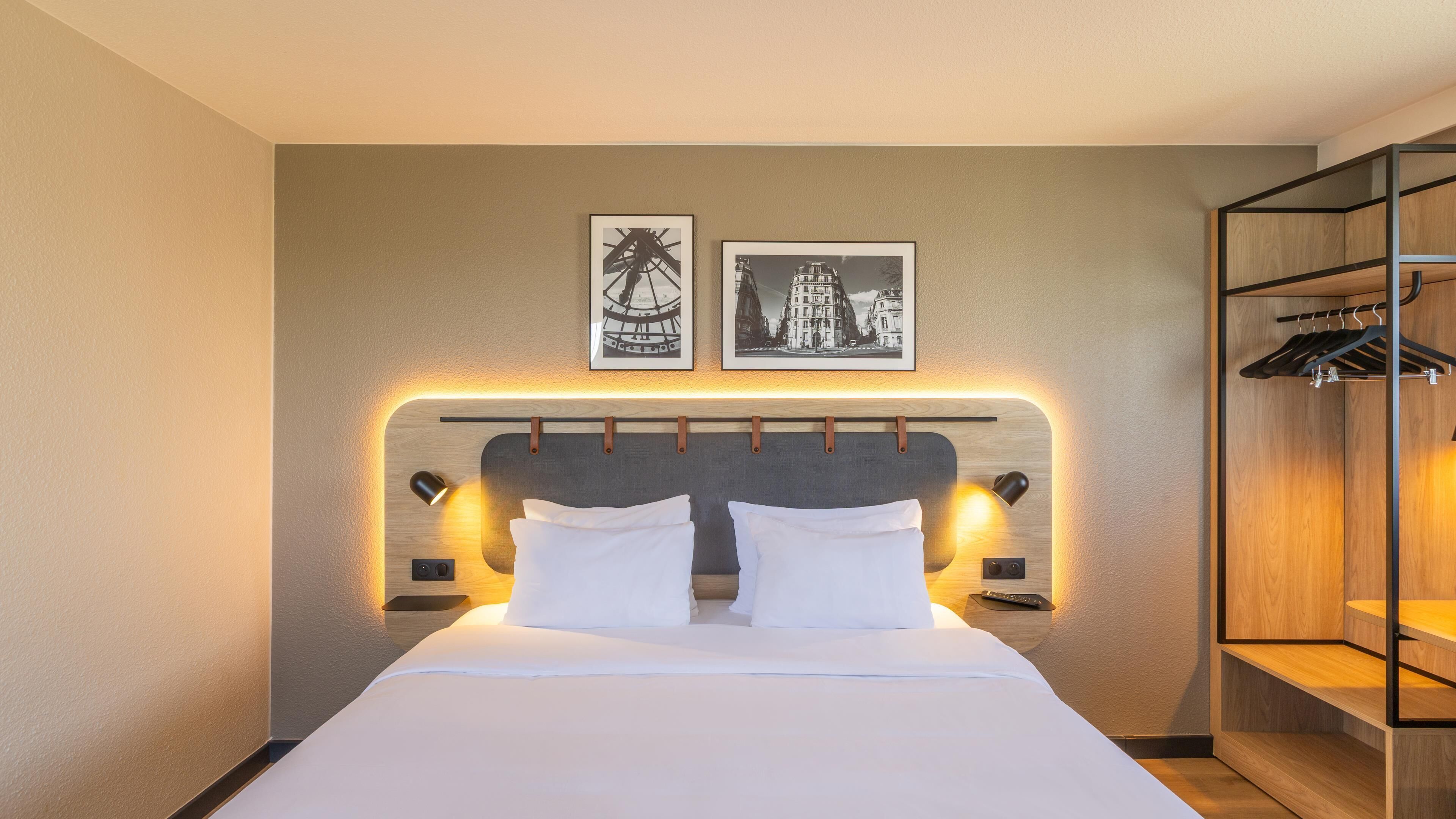 Habitación de hotel con cama, dos cuadros en la pared, cabecero con iluminación integrada y armario de madera con espacio para colgar ropa.