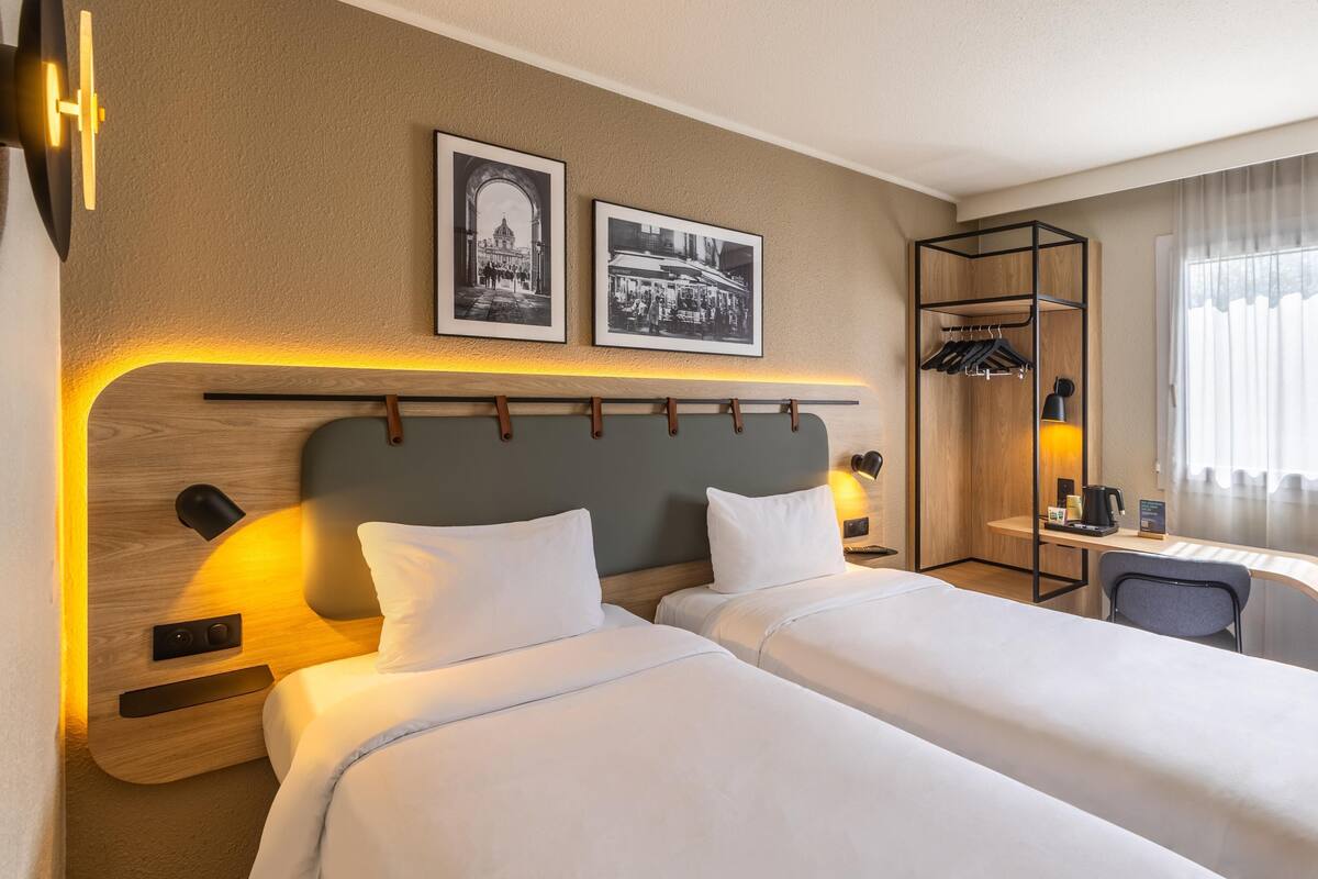 A hotel room with two beds, a desk, a chair, a coat rack, and two framed pictures on the wall.