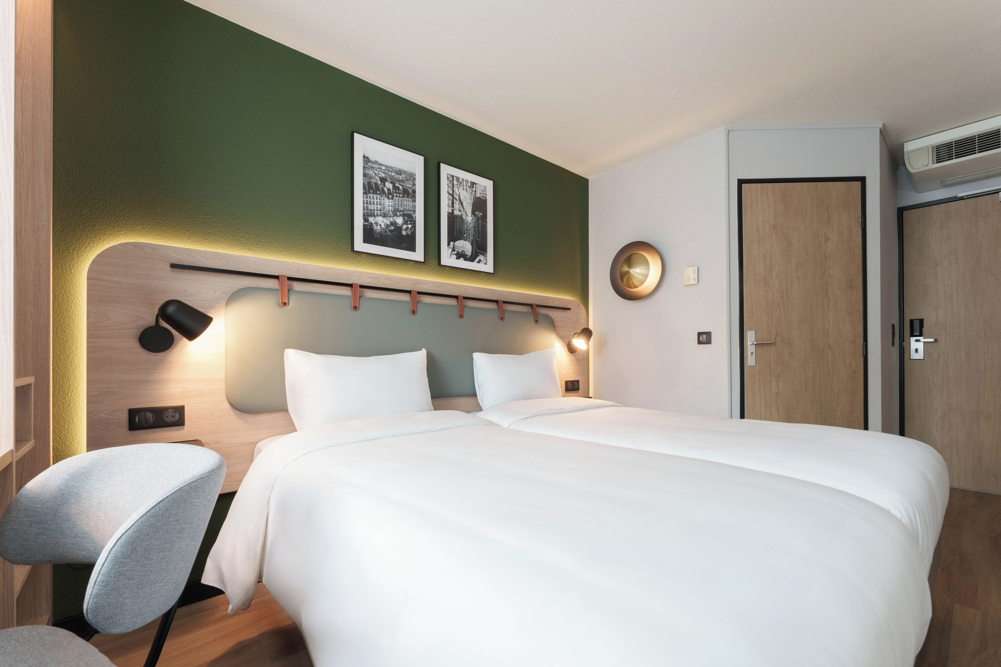 A modern hotel room with a large bed, a seating area with a chair, a wall-mounted shelf, and two framed pictures.