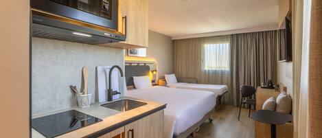 A hotel room with a kitchenette, two beds, a desk, and a chair.