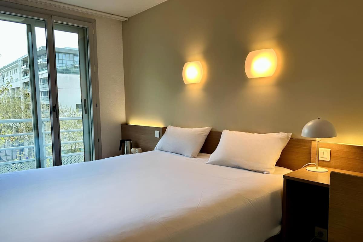 Una habitación de hotel con una cama, dos lámparas de noche, una mesita de noche y vistas a un edificio a través de un ventanal grande.