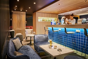 A modern bar area with a bar counter, seating with blue upholstered chairs, and a barista preparing a drink.