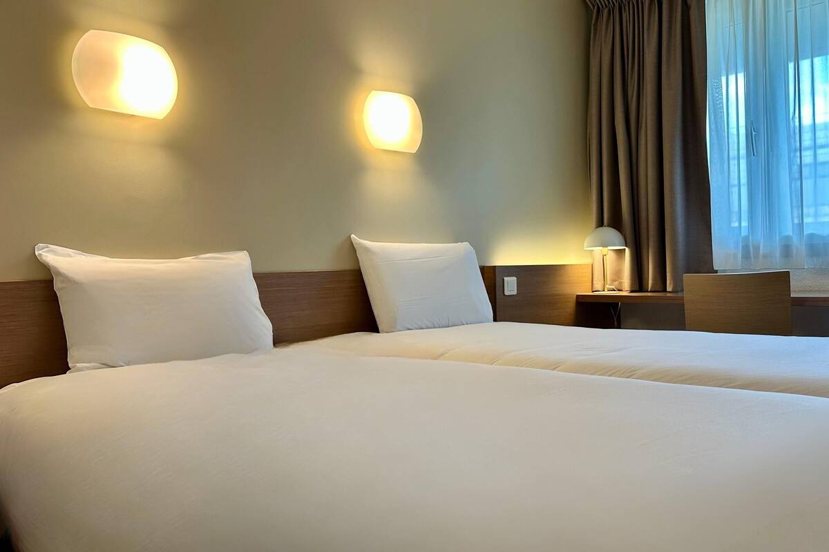 Una habitación de hotel con dos camas, una mesita de noche con lámpara y una ventana con cortinas.