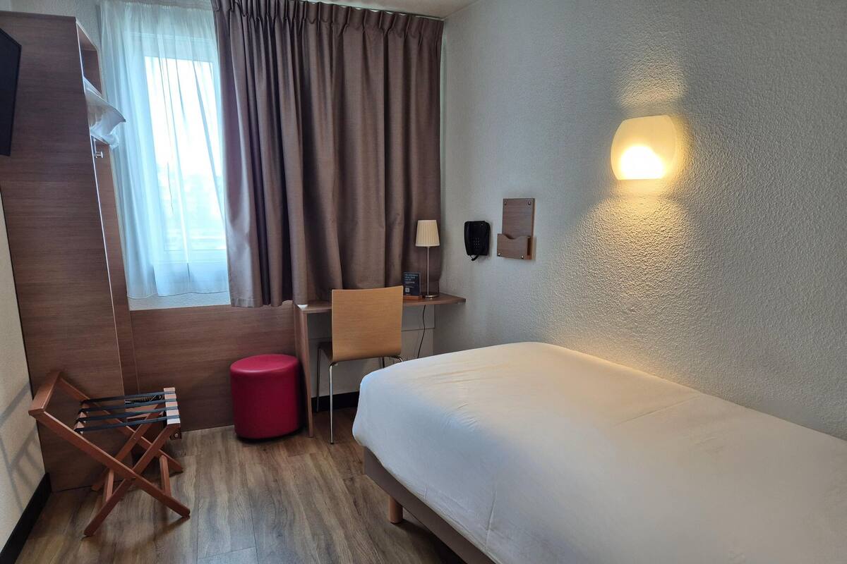 Habitación de hotel con cama, escritorio, silla, reposapiés rojo, lámpara y teléfono en la pared.