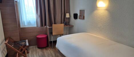 A hotel room with a bed, a desk, a chair, a red ottoman, a lamp, and a telephone on the wall.