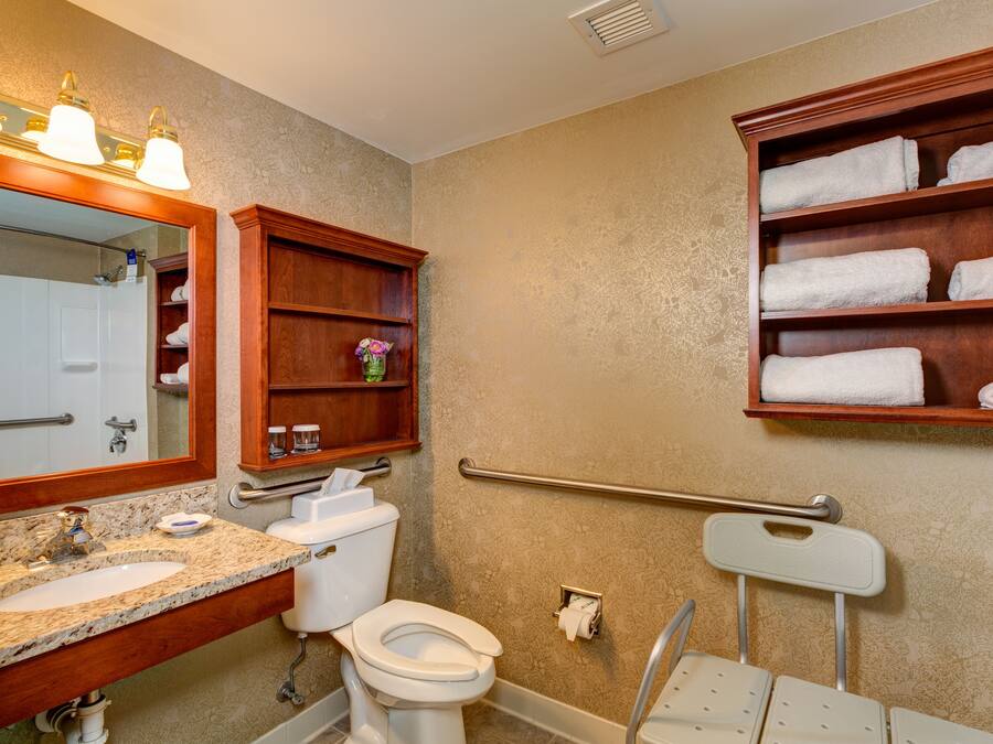 Une salle de bain avec un lavabo en marbre, des toilettes, un miroir avec éclairage intégré, une étagère avec des serviettes et une barre d’appui.