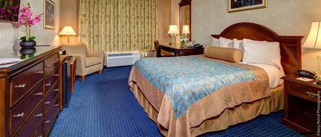 A hotel room with a large bed, a desk with a chair, a dresser, a mirror, and a window with curtains.