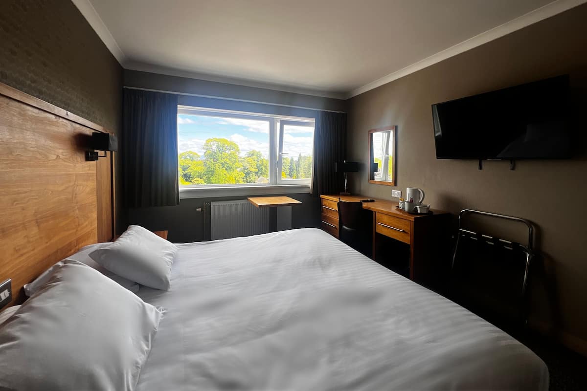 Ein Hotelzimmer mit einem großen Bett, einem Schreibtisch, einem Sessel, einem Flachbildfernseher und einem Fenster mit Blick auf Bäume.