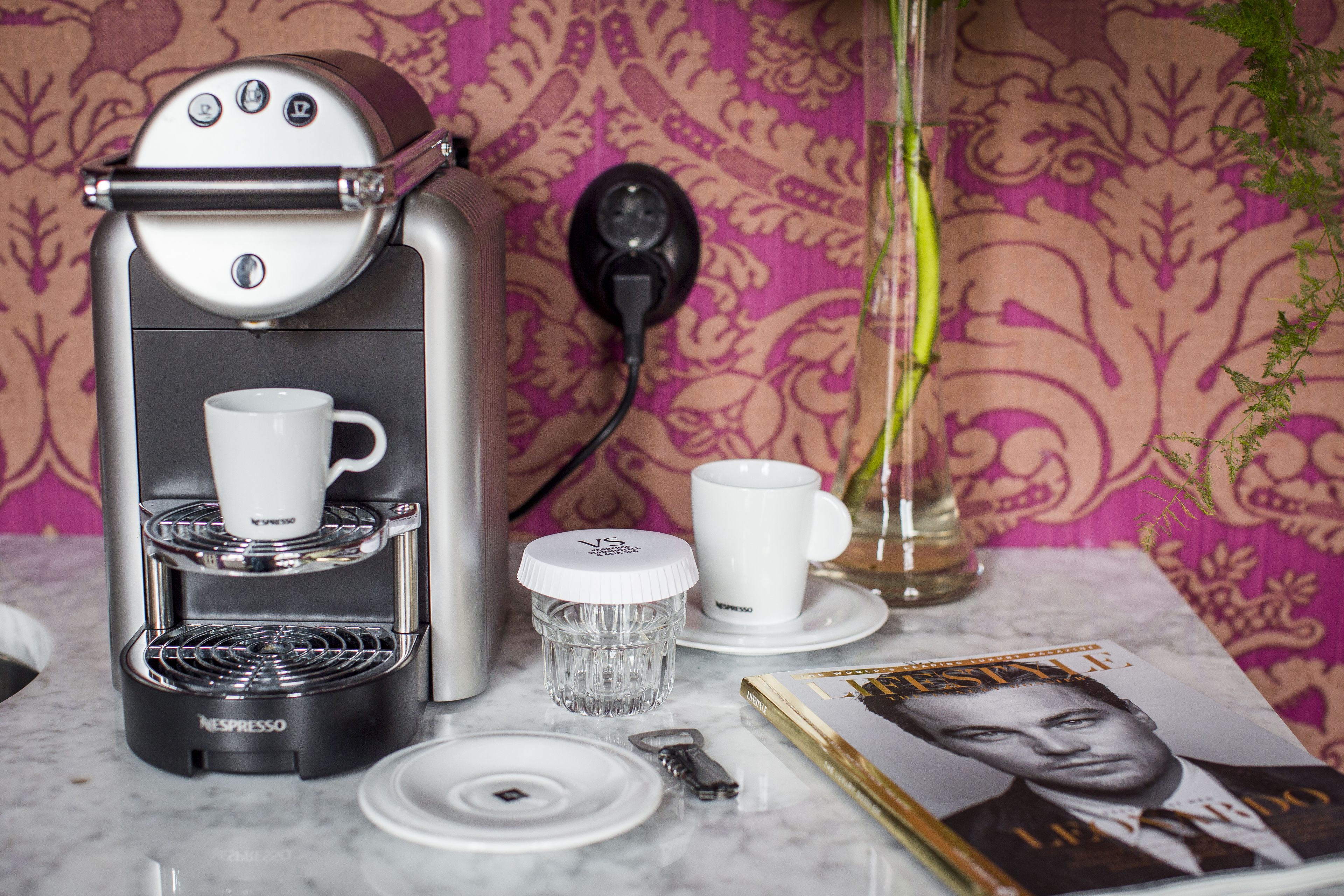 A Nespresso coffee machine, two white cups, a glass, a key, and a magazine on a table with a patterned wallpaper background.