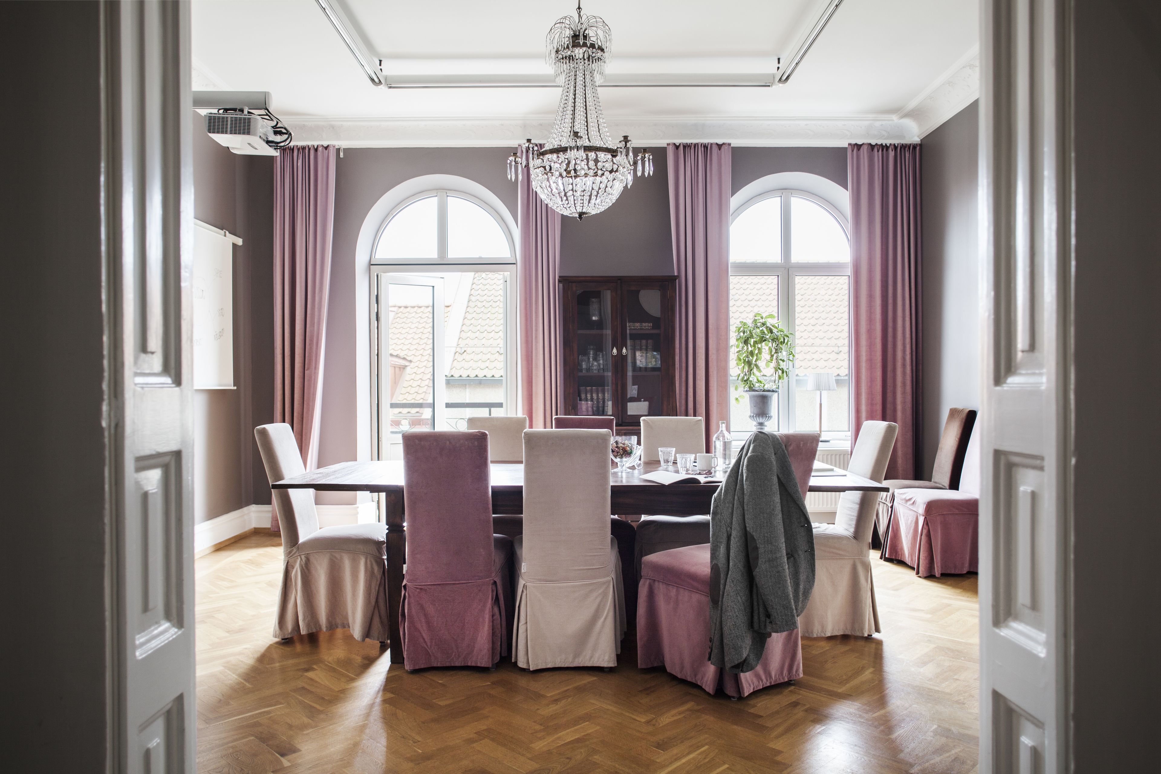 A dining area with a wooden herringbone floor, a chandelier, and a large window with purple curtains.