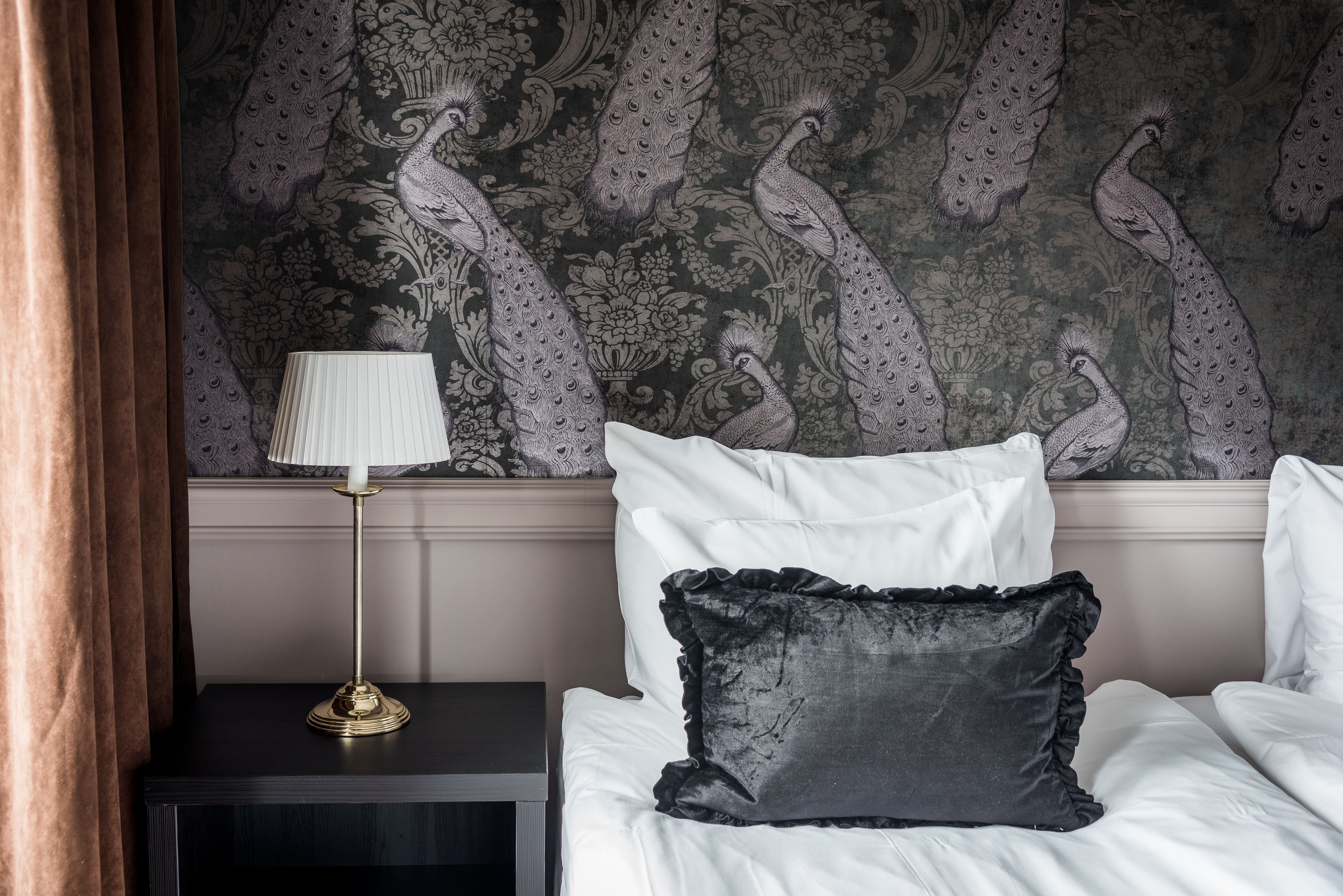 Un lit bien fait, avec du linge de lit blanc et un oreiller noir à volants, une lampe à abat-jour doré posée sur une table de chevet sombre, et un mur orné d’un motif de paon.
