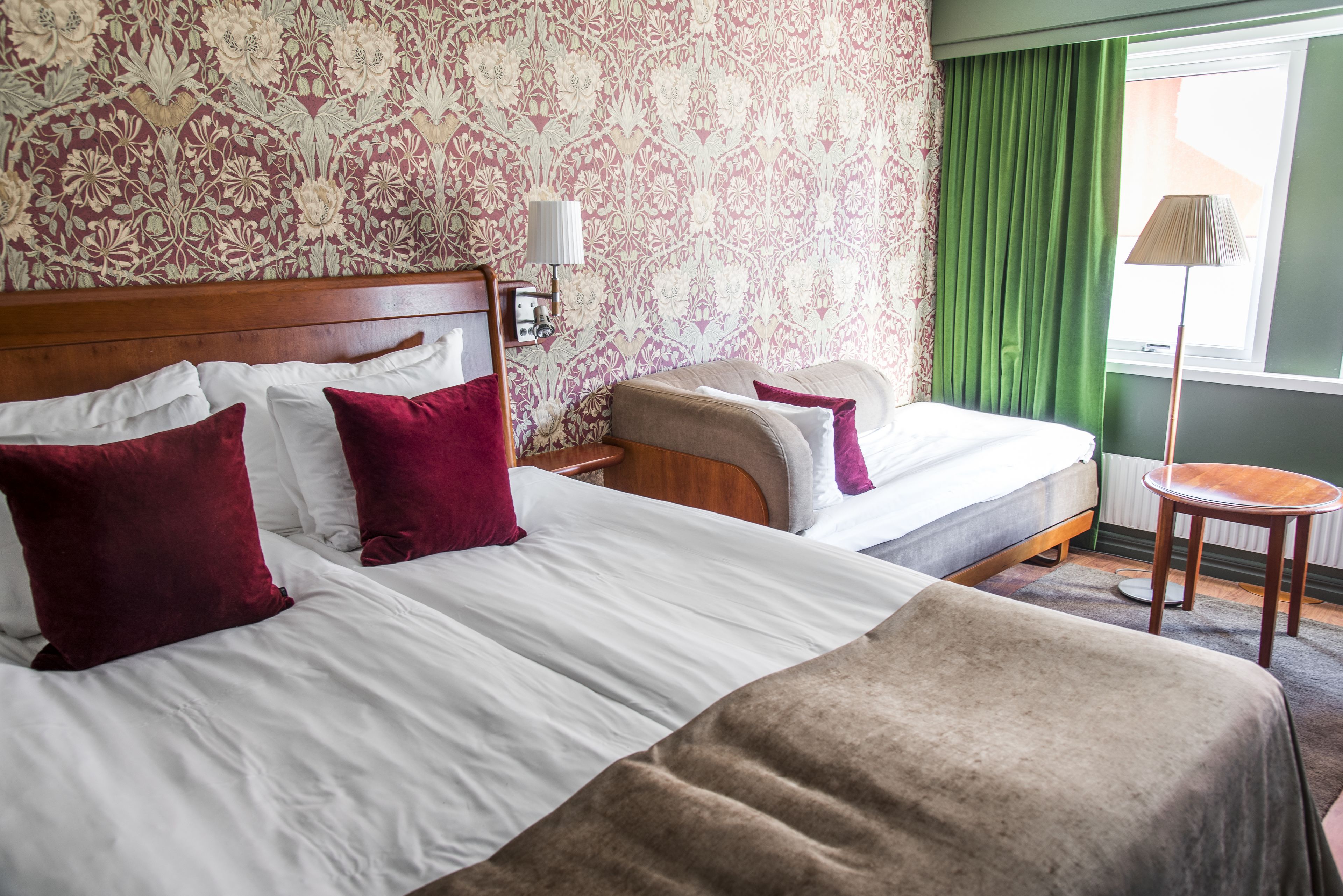 Une chambre d’hôtel avec deux lits, une tête de lit en bois, une table de chevet, une lampe et une vue sur l’extérieur par une fenêtre ornée de rideaux verts.