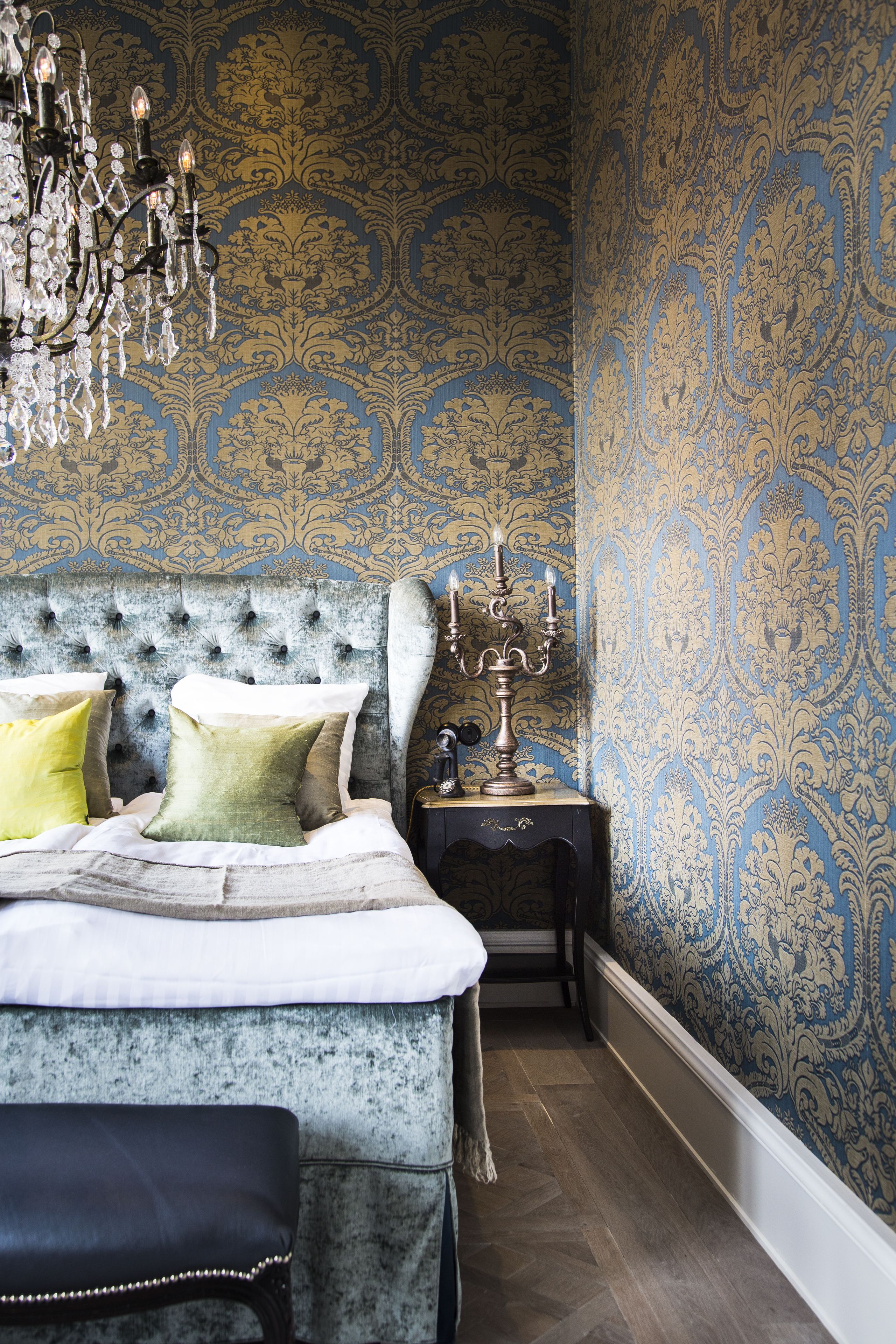 A bedroom with a tufted headboard, a chandelier, patterned wallpaper, and a nightstand with a candelabra.
