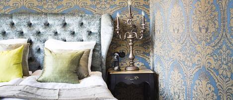 A bedroom with a tufted headboard, a chandelier, patterned wallpaper, and a nightstand with a candelabra.