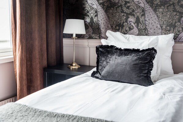 A neatly made bed with white linens and a black pillow, a gold lamp on a bedside table, and a patterned wallpaper with peacock designs. - Varbergs Stadshotell & Asia Spa (Varberg)