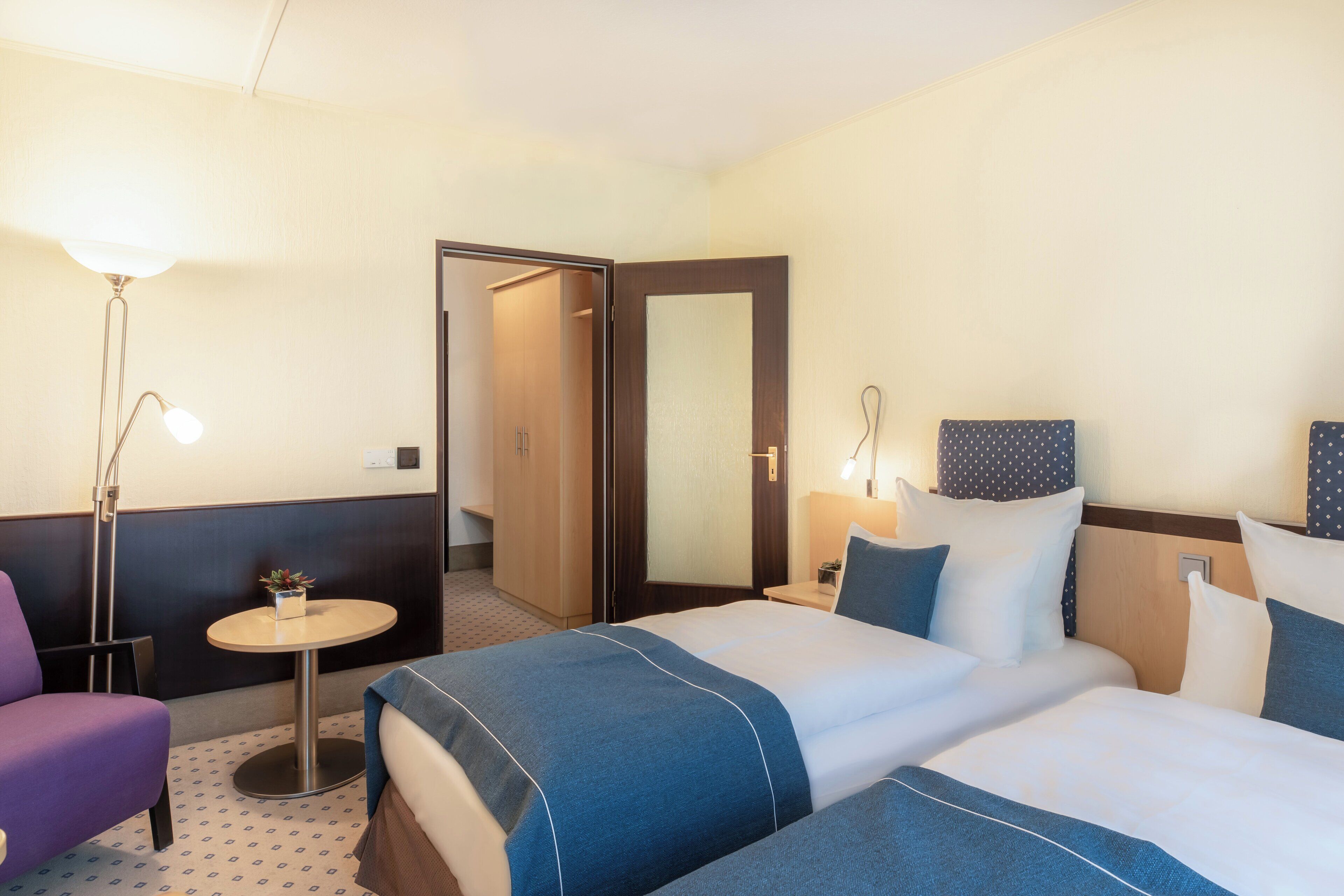 Ein Hotelzimmer mit zwei Betten, einem kleinen runden Tisch, einem Stuhl, einer Lampe und einer Tür zu einem anderen Raum.