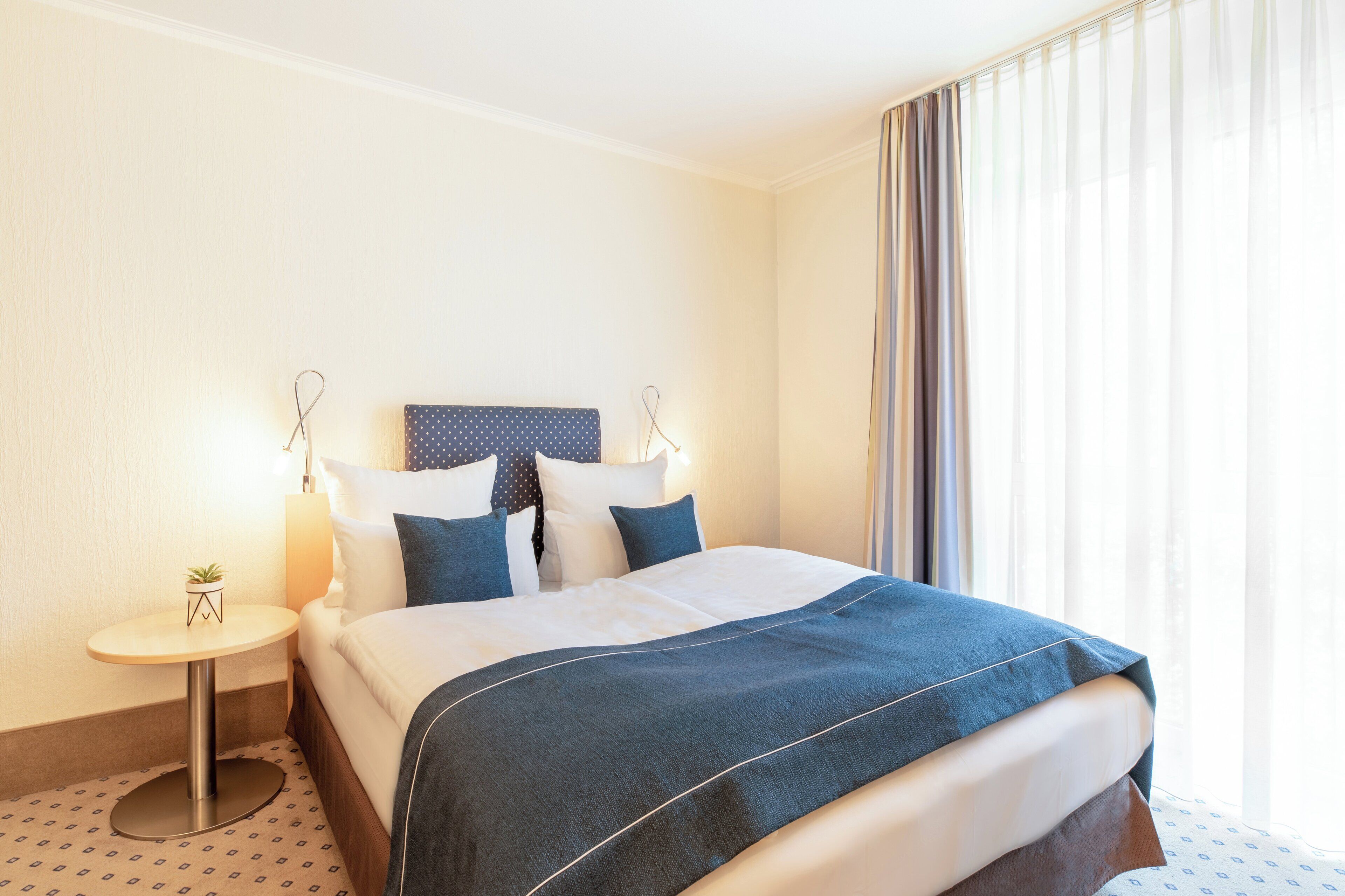 A hotel room with a neatly made bed, a small side table with a lamp, and a window with sheer curtains.