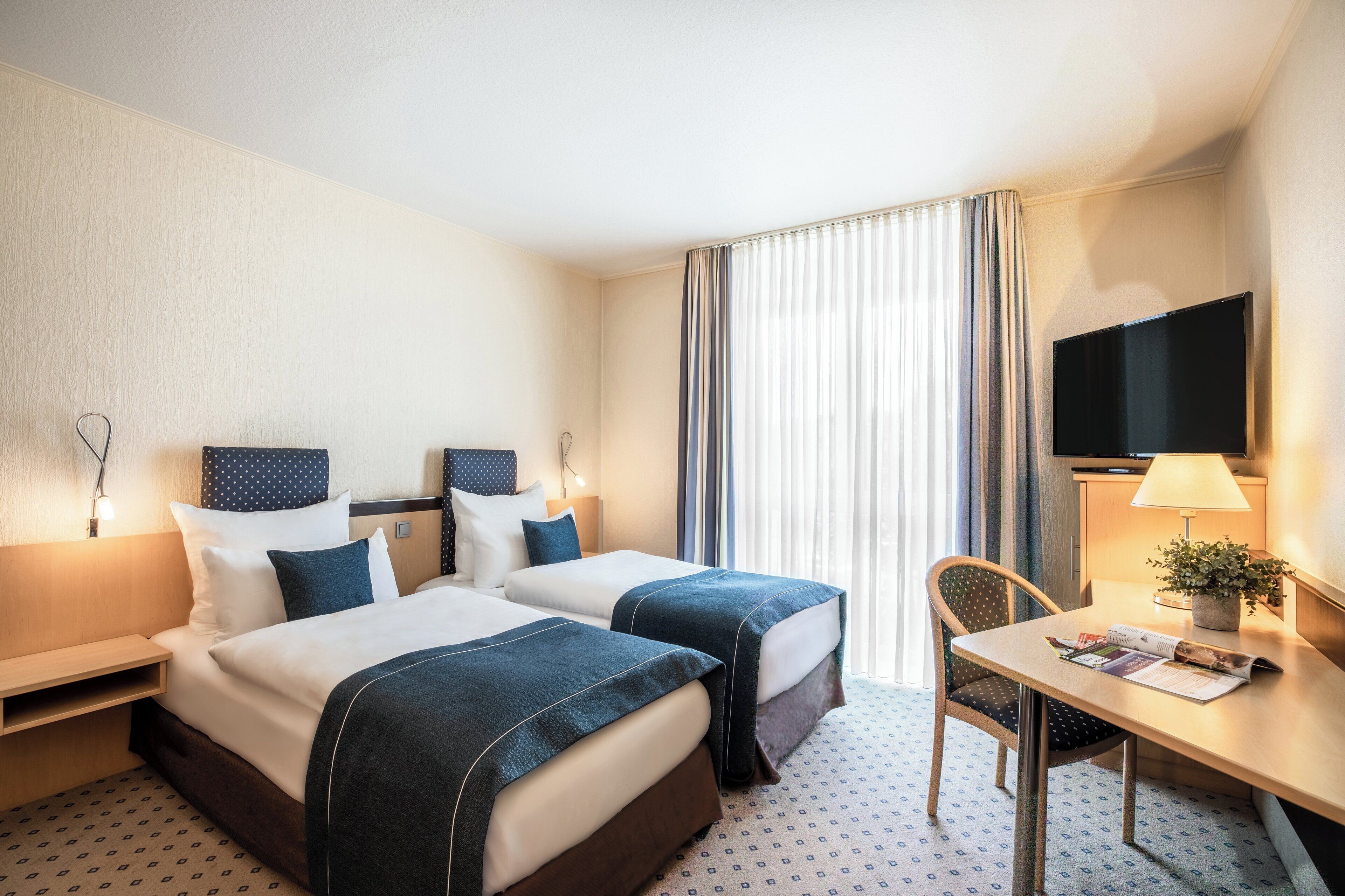 Habitación de hotel con dos camas, un escritorio, una silla, un televisor y una ventana con cortinas.