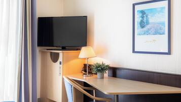 A hotel room with a flat-screen TV mounted on the wall, a bedside table with a lamp and a potted plant, a framed picture on the wall, and a window with curtains.