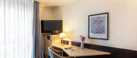 Habitación de hotel con cama, escritorio con silla, televisor, lámpara y un cuadro en la pared.