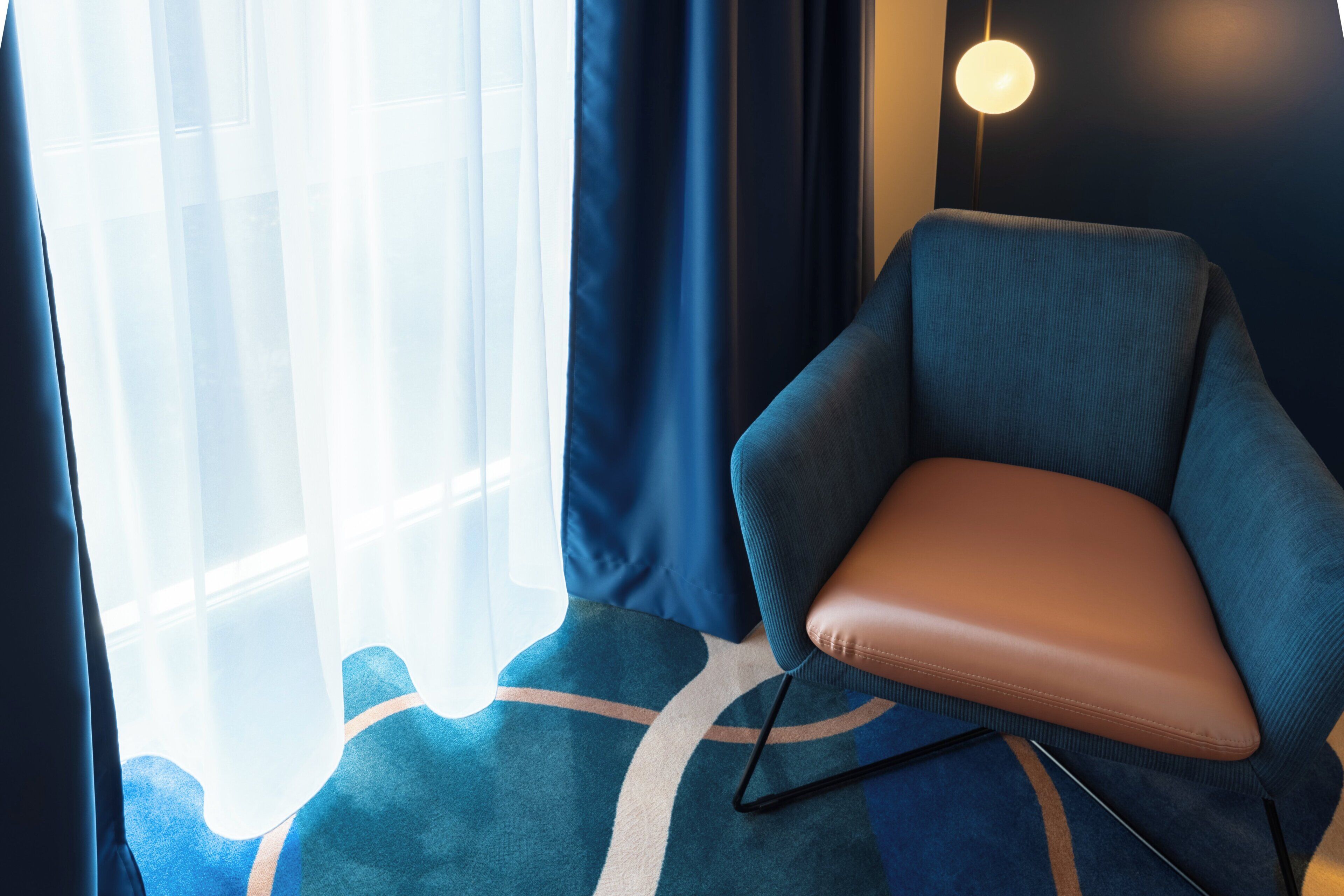 Ein modernes Zimmer mit blauem Ohrensessel, Teppich mit Muster und einem großen Fenster mit leichtem Vorhang.