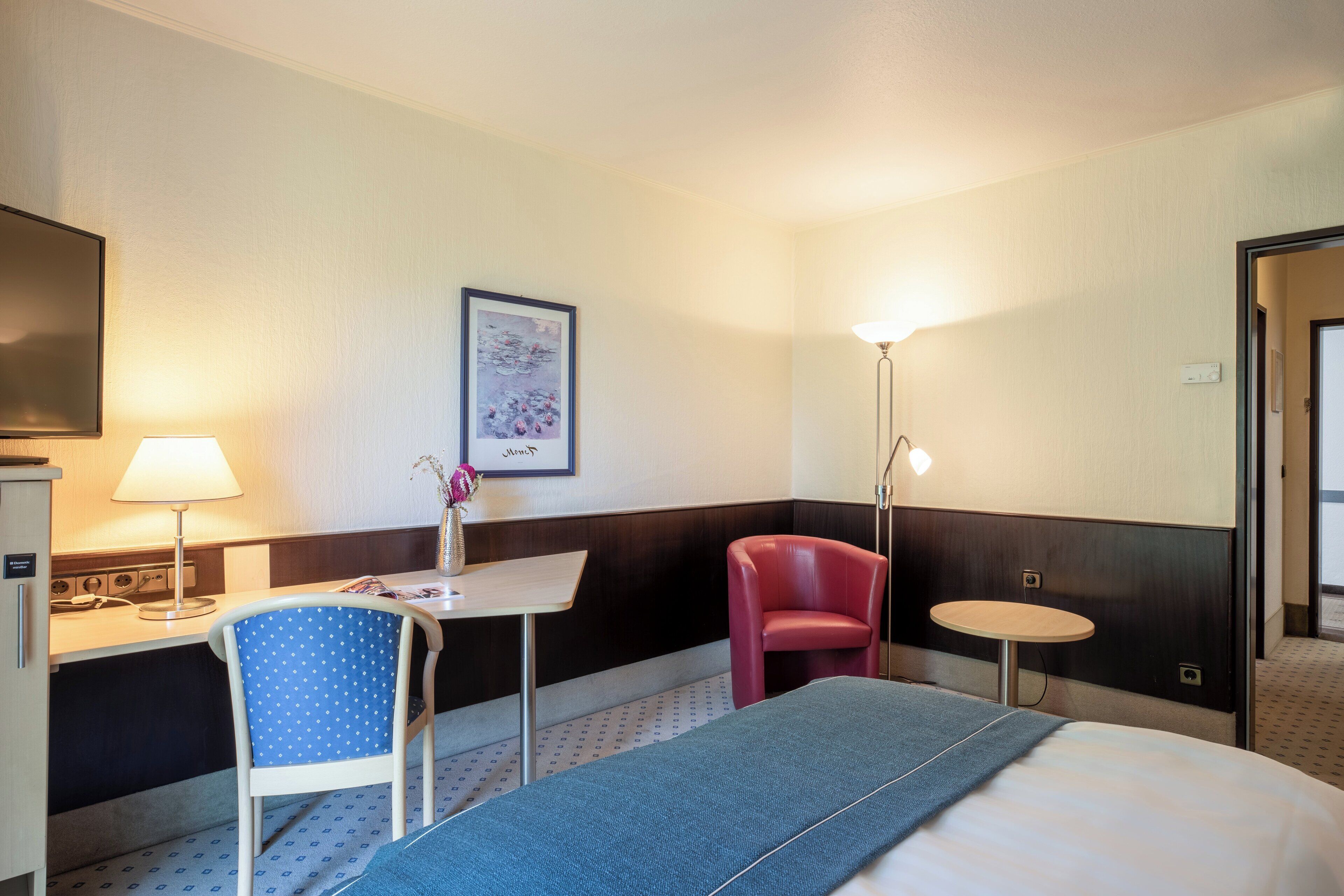 A hotel room with a bed, a desk, a chair, a small table, and a lamp.