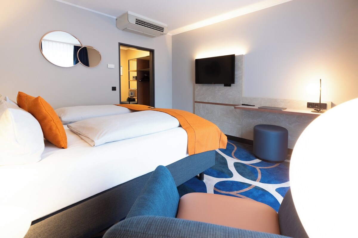 Une chambre d’hôtel avec un lit, un téléviseur fixé au mur, une table de chevet et une salle de bain visible à travers une porte ouverte.