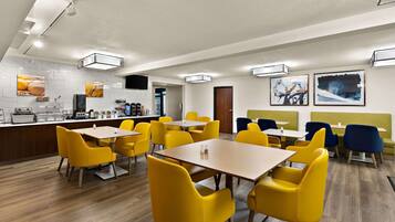 A modern dining area with yellow chairs and tables, a kitchen in the background, and artwork on the walls.