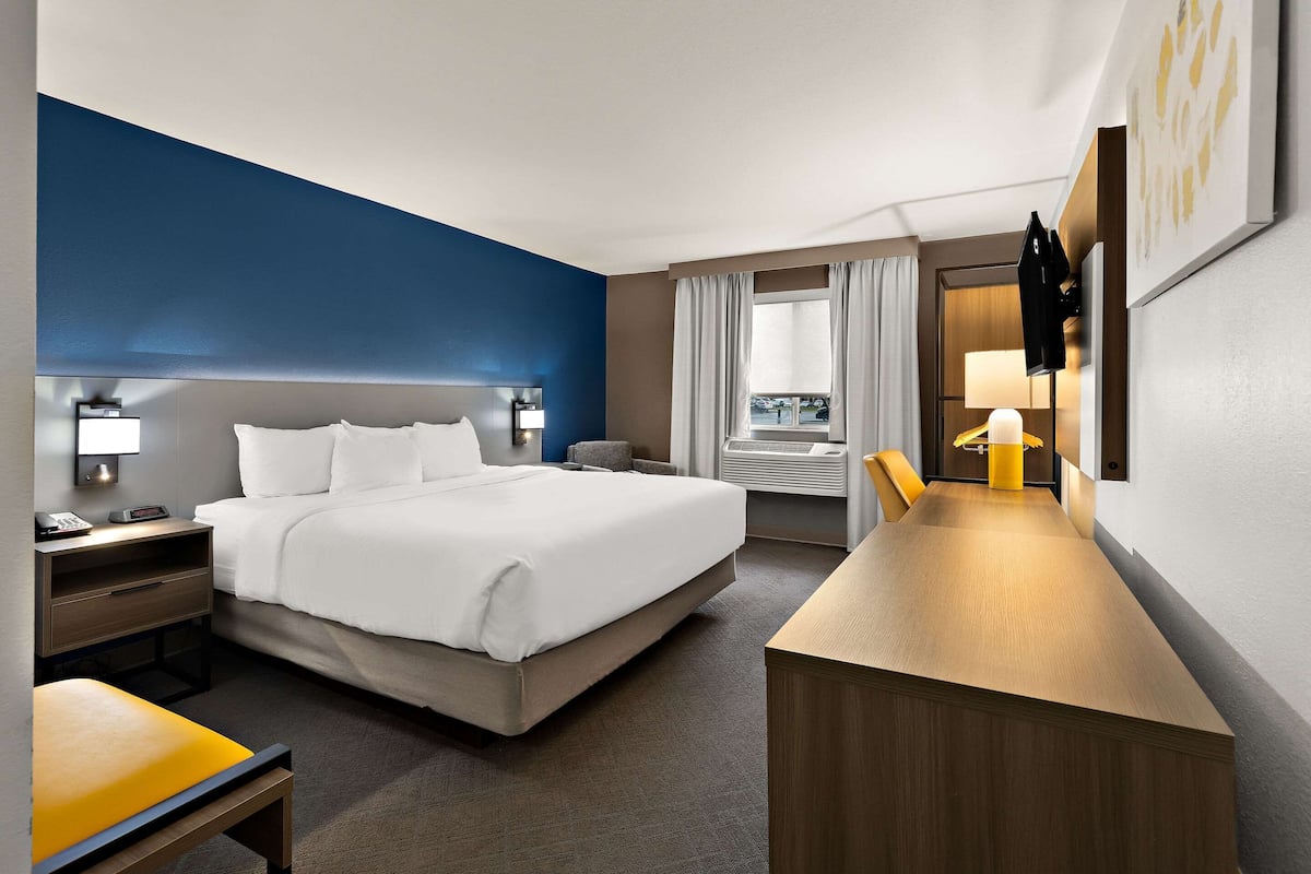 Une chambre d’hôtel comprenant un lit, une table de chevet, une lampe, un bureau, une chaise et une fenêtre avec des rideaux.