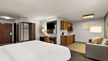 A hotel room with a large bed, a desk with a chair, a kitchenette, a sofa, and a TV.