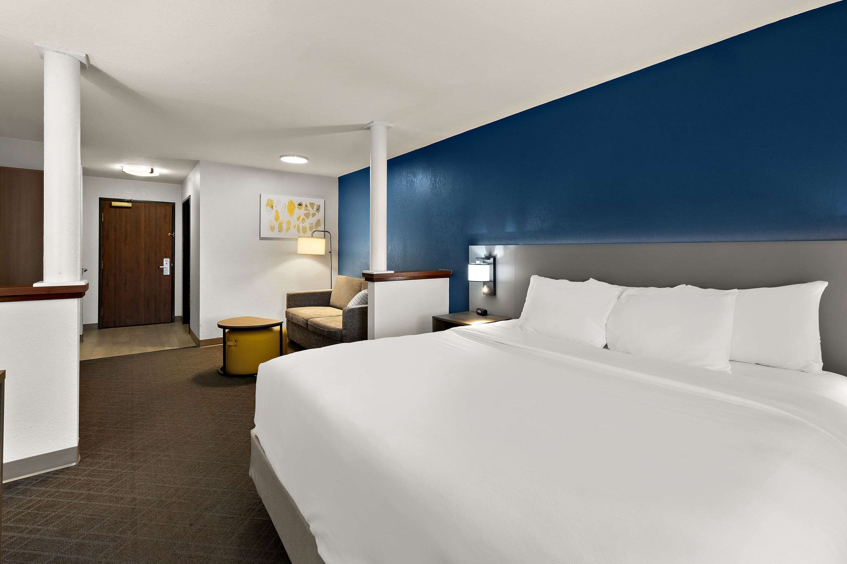 A hotel room with a large bed, a desk, a chair, a lamp, and a door.