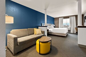 Habitación de hotel con pared azul decorativa, un sofá, una cama, un escritorio y un reposapiés redondo amarillo.