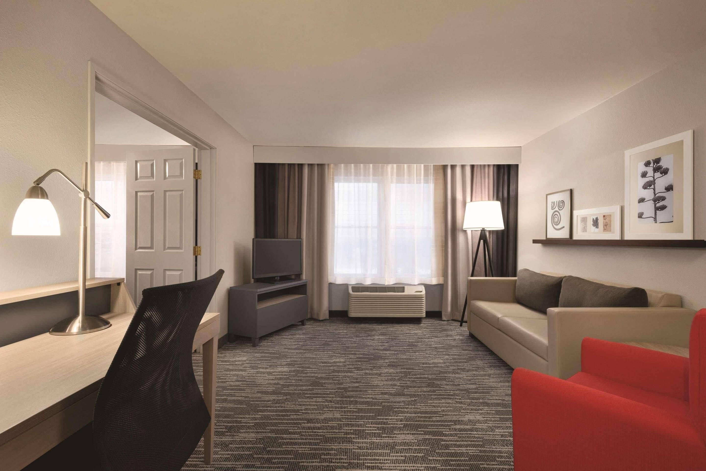 Une chambre d’hôtel équipée d’un bureau, d’une télévision, d’un fauteuil rouge, d’un canapé et d’une grande fenêtre avec des rideaux.