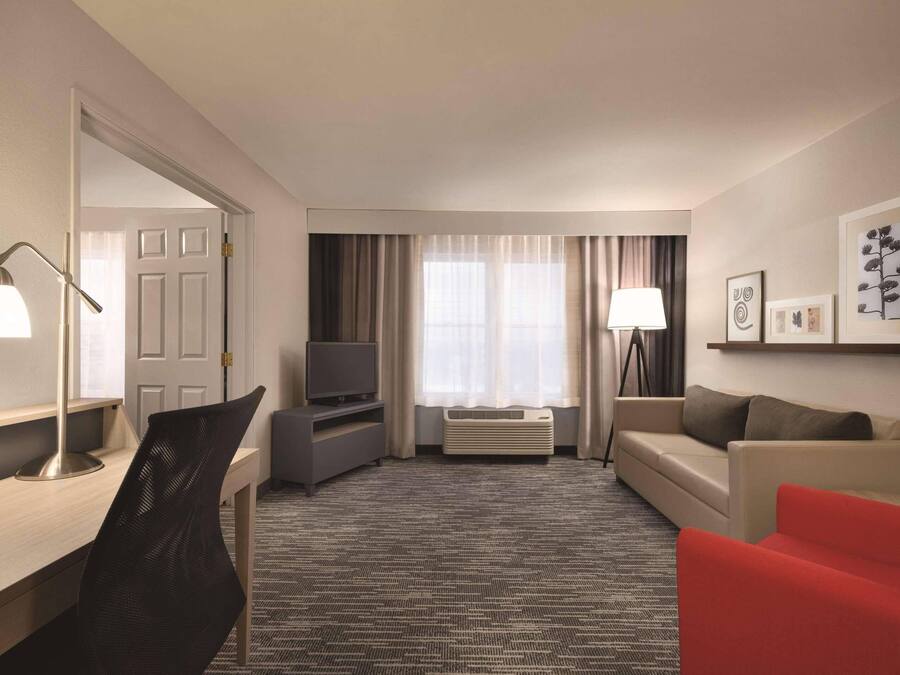 Habitación de hotel con escritorio, televisor, una silla roja, sofá y un ventanal con cortinas.