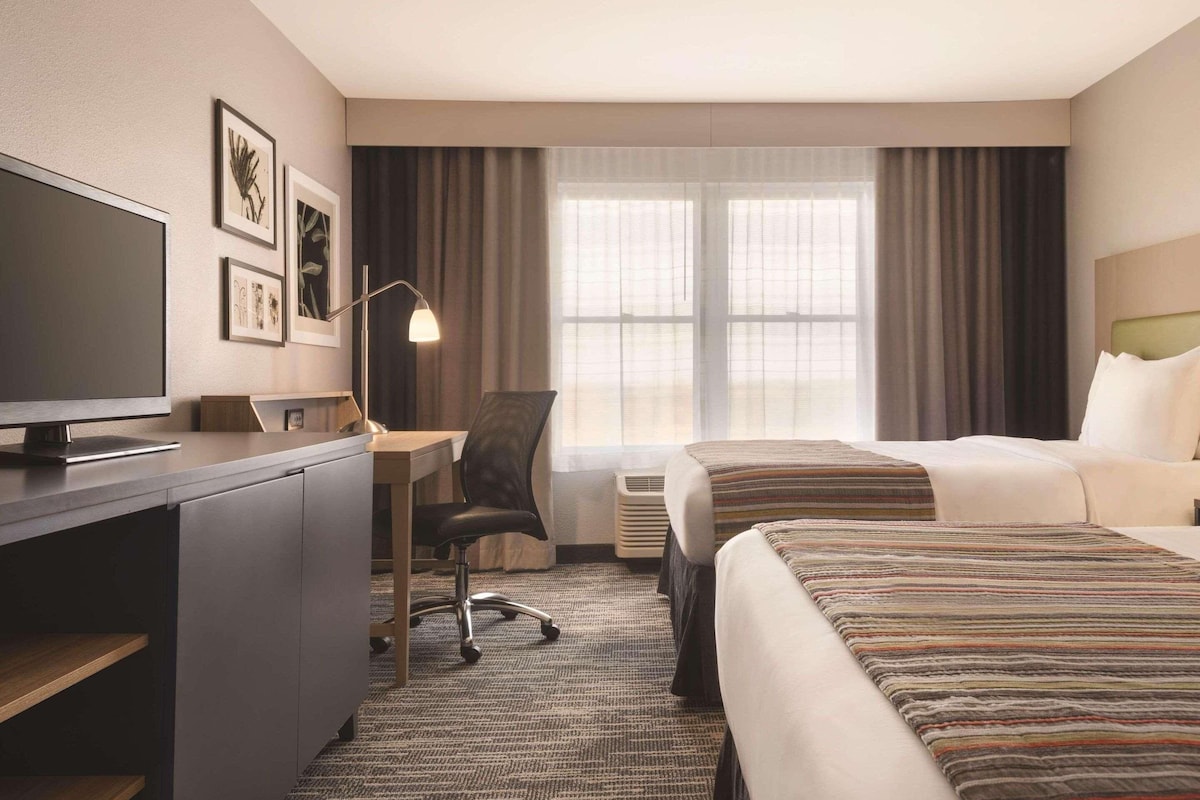 A hotel room with a desk, chair, two beds, a TV, and a large window with curtains.