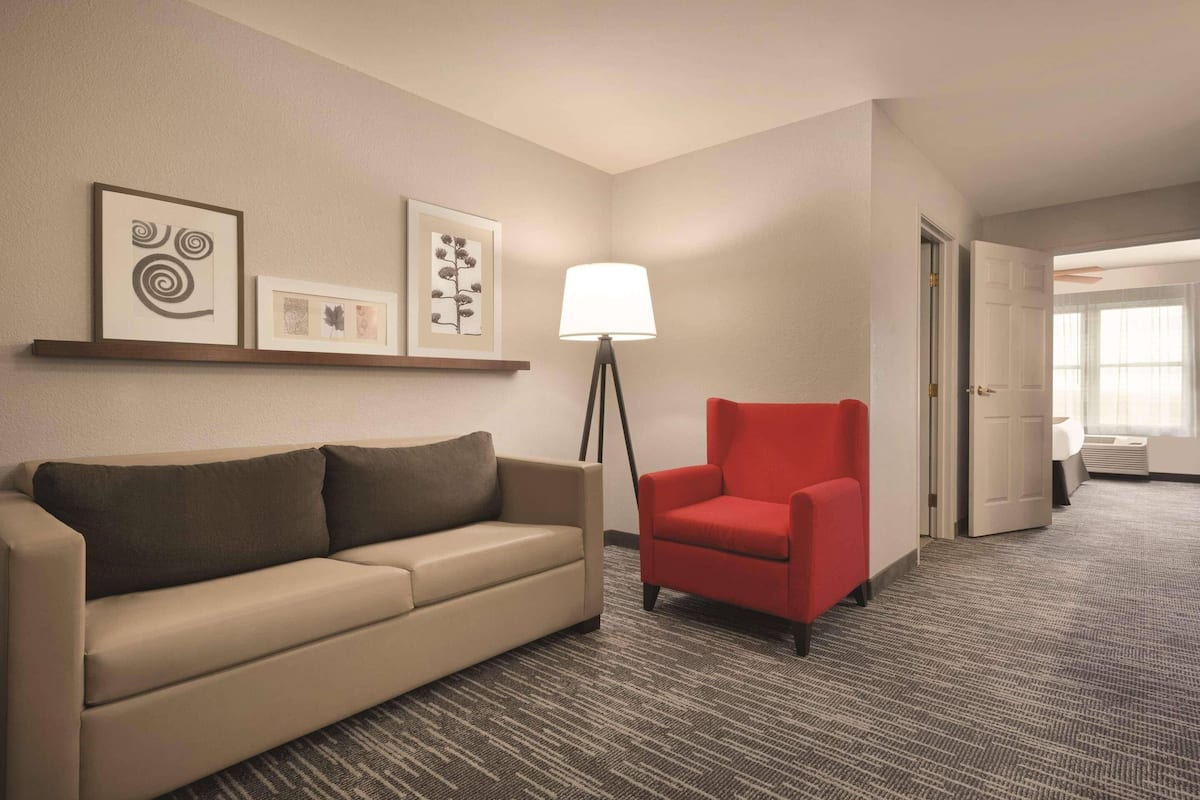 Une chambre d’h ôtel avec un canapé beige, un fauteuil rouge, une lampe sur pied et des œuvres d’art encadrées accrochées au mur.