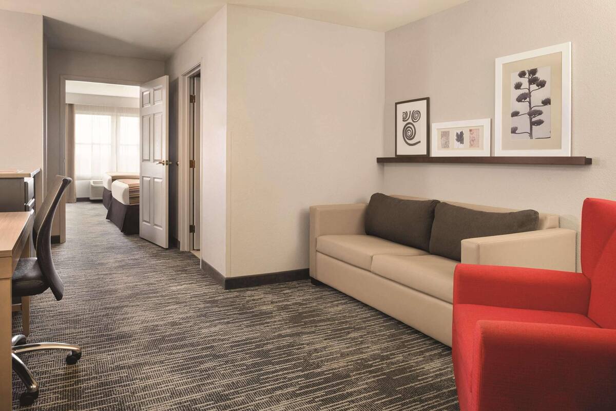 Une chambre d’hôtel avec un sol recouvert de moquette, un canapé beige, un fauteuil rouge, un bureau avec une chaise et un lit à l’arrière-plan.