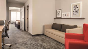 Habitación de hotel con piso alfombrado, un sofá color beige, un sillón rojo, un escritorio con silla, y una cama al fondo.
