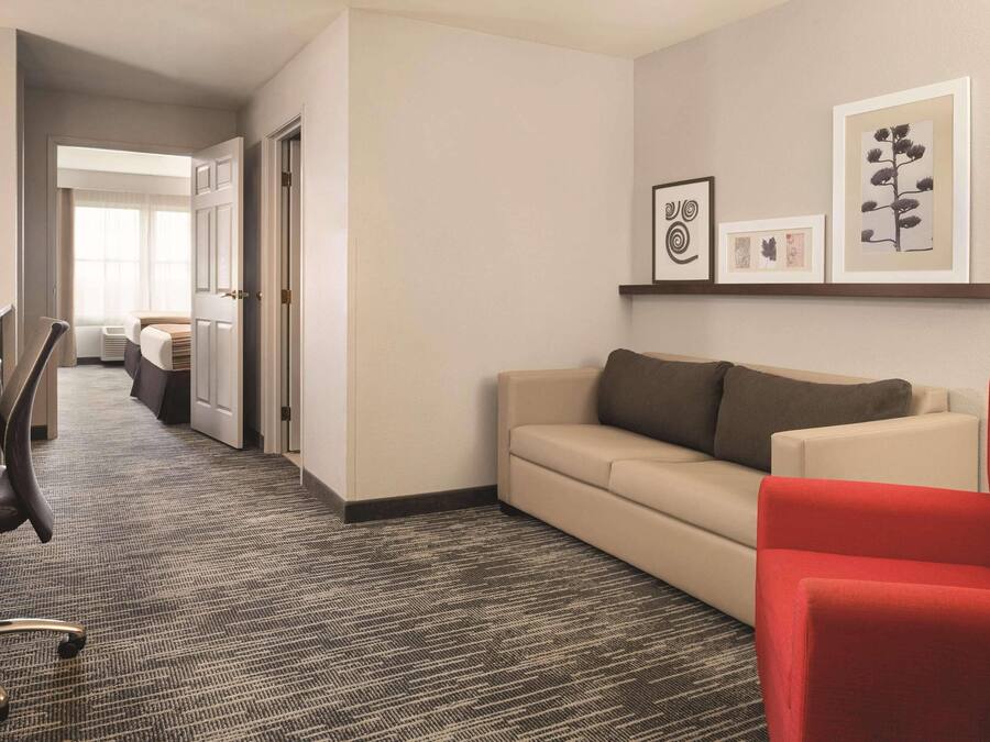 Habitación de hotel con piso alfombrado, un sofá color beige, un sillón rojo, un escritorio con silla, y una cama al fondo.