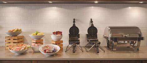 Una cocina con salpicadero de azulejos blancos, armarios de madera y una encimera con planchas para gofres, una parrilla y tazones con comida.