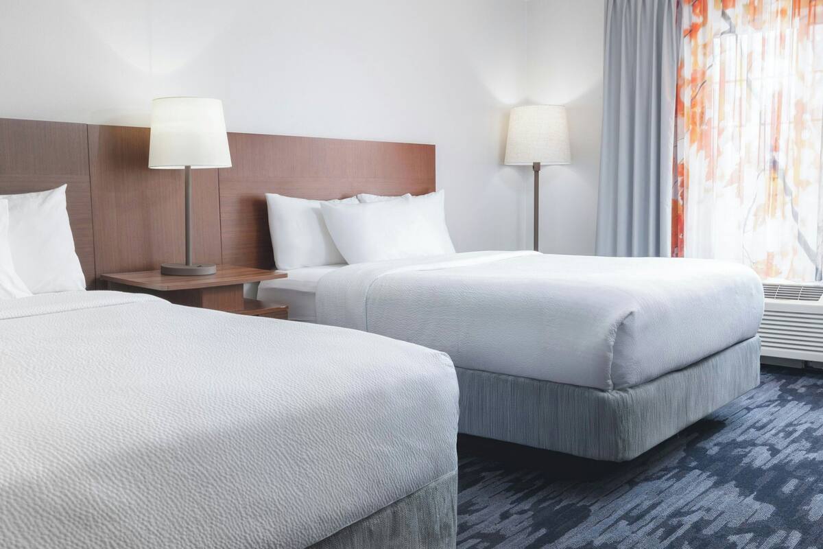 Une chambre d’hôtel avec deux lits, une table de chevet, une lampe et une fenêtre avec des rideaux.