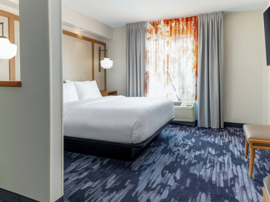 A hotel room with a large bed, a wooden headboard, a bedside table, a chair, a window with curtains, and a patterned carpet.