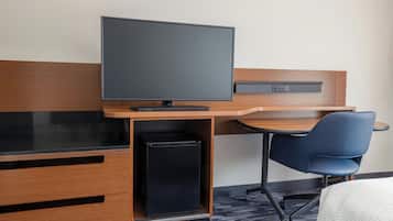 Habitación de hotel con escritorio de madera, televisor de pantalla plana, refrigerador negro compacto, silla azul y cama con cubierta blanca.