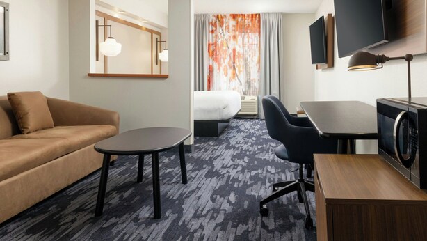 A hotel room with a sofa, a small round table, a desk with a chair, a bed, a TV, and a large window with curtains. - Fairfield Inn & Suites by Marriott Cincinnati Eastgate (Cincinnati)