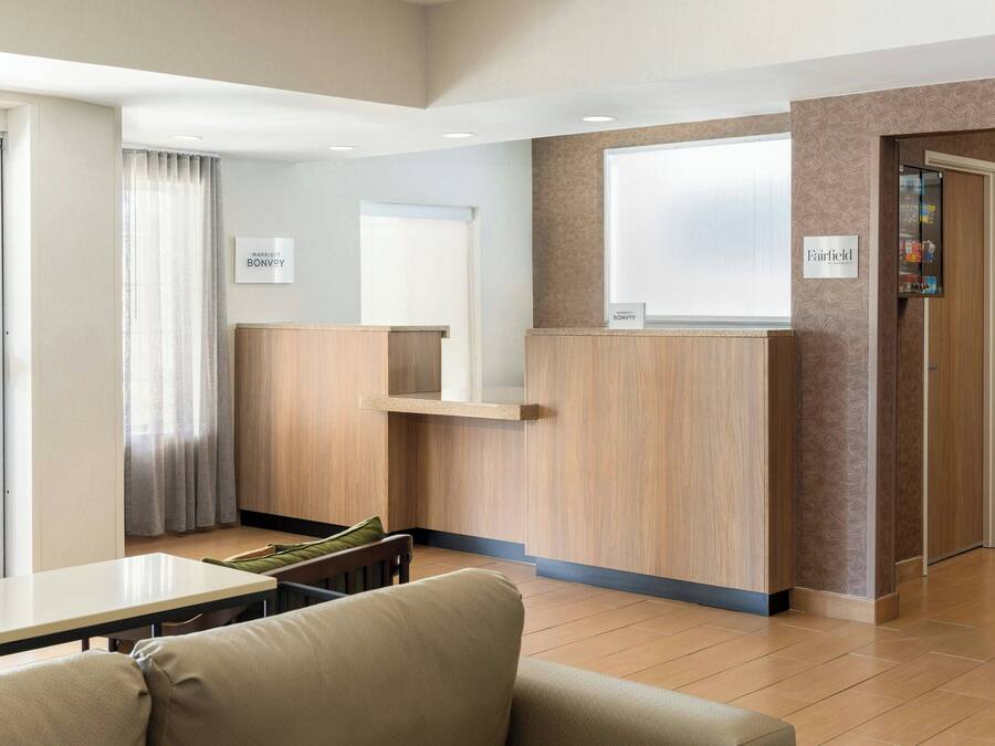 Uma área de recepção de hotel com balcão, sala de estar com sofá e mesa, e uma placa na parede com o nome do hotel.