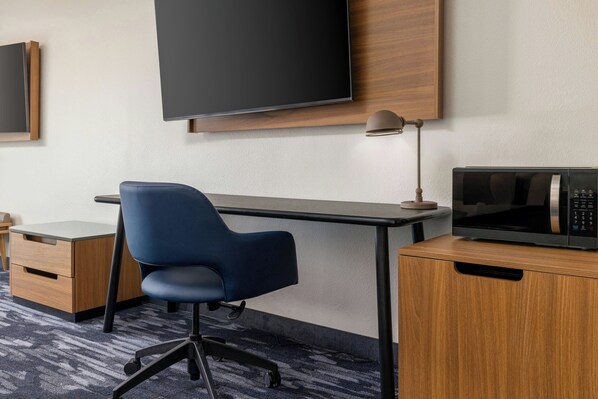 A hotel room with a desk, a blue office chair, a flat-screen TV mounted on the wall, a microwave on the desk, and a wooden cabinet.